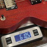 A close-up of a red electric guitar body resting on a digital scale displaying 6 lbs 10.5 oz, with the guitar pickups and switch visible.