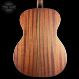 The back of an acoustic guitar made of rich, warm brown wood with prominent grain patterns, shown against a solid black background with a Tone Shop watermark in the top left corner.