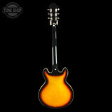 A classic double-cutaway electric guitar shown from the back, featuring a vibrant sunburst finish that transitions from bright orange in the center to dark black around the edges. The headstock is black with silver tuning pegs, set against a solid black background.