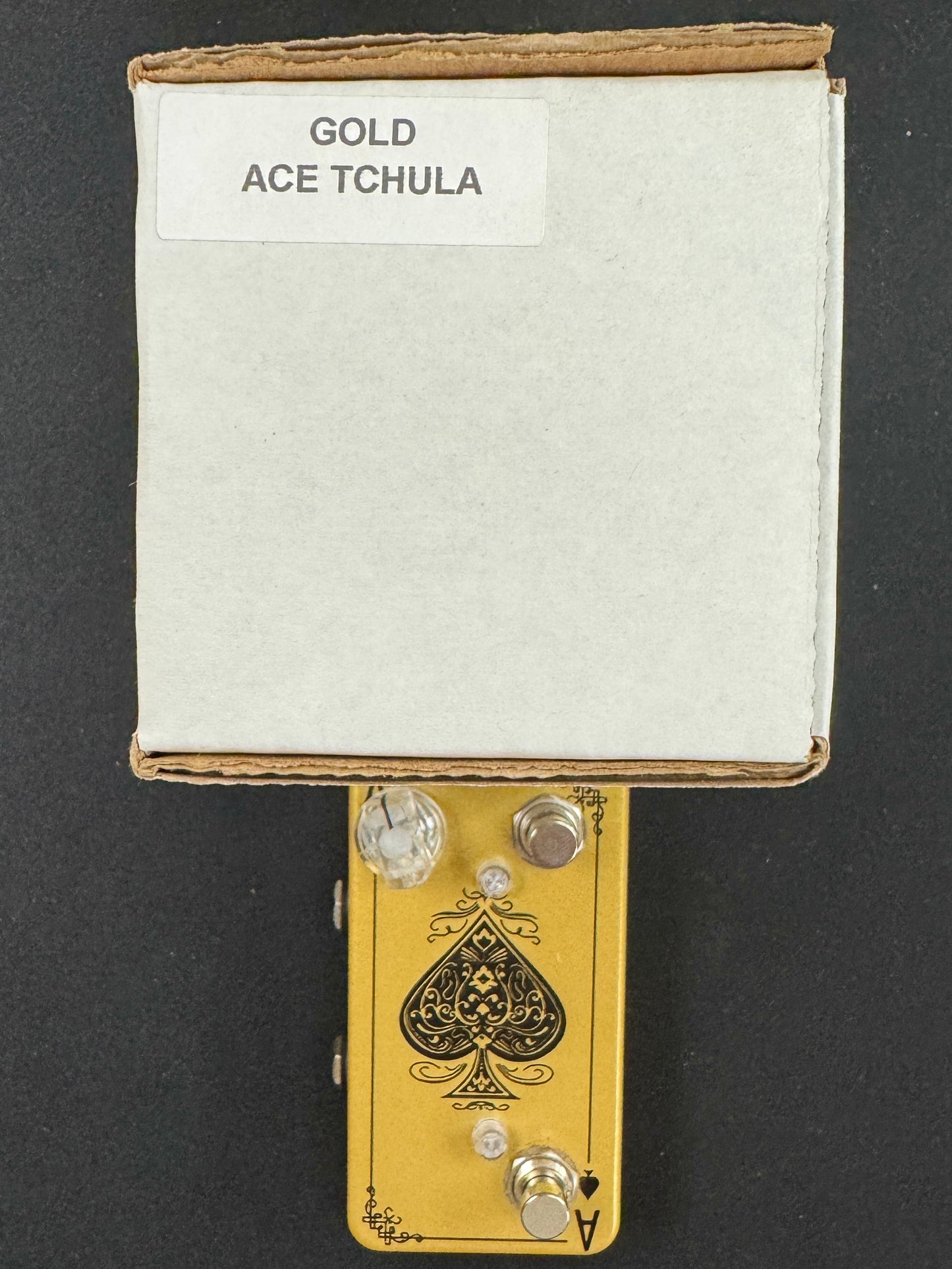 Gold Ace Tchula guitar effects pedal with packaging on a black background