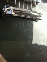 A close-up shot of a chrome guitar tailpiece assembly attached to a glossy black guitar body, showing some surface scratches and reflections.