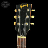Close-up of a Gibson acoustic guitar headstock against a black background, showing the Gibson logo in gold, six tuning pegs, and part of the dark wood fretboard.