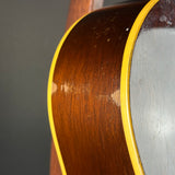 Close-up of the side of an acoustic guitar showing the dark brown wood body, a bright yellow binding edge, and part of the silver metallic backplate.
