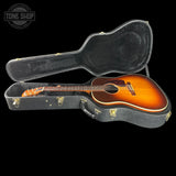 An acoustic guitar with a sunburst finish displayed inside an open black hard case against a solid black background. A Tone Shop watermark is visible in the top-left corner.