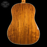 The back of an acoustic guitar made of rich, warm brown wood with prominent grain patterns, shown against a solid black background with a Tone Shop watermark in the top left corner.