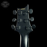 Close-up of the headstock of a black electric guitar against a black background, showing six tuning pegs and a gold serial number. A Tone Shop watermark is visible in the top left corner.
