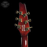 Close-up of the headstock of a guitar with a glossy dark red finish and gold tuning pegs, shown against a solid black background. A Tone Shop watermark is visible in the top left corner.