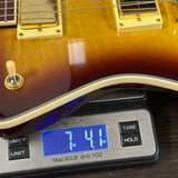 A close-up of a guitar body resting on a digital scale, showing a sunburst finish with cream trim and pickups. The scale displays a weight of 7.