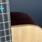 Close-up detail of an acoustic guitar showing the dark brown wooden top edge, the light natural wood body, and part of the black fretboard with gold strings against a dark gray background.