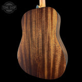 Close-up of the back of an acoustic guitar showing a rich, warm brown wood grain finish, set against a solid black background with a Tone Shop watermark in the top left corner.