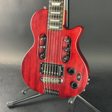 A red electric guitar with a glossy cherry finish and black hardware, shown from the side on a stand against a dark gray background.