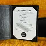 A Gibson Custom Certificate of Authenticity insert displayed inside an open black presentation folder on a textured tan surface. The card shows product details for an LP Custom Q guitar finish and includes the Gibson logo and a Dealer Select logo.