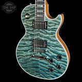 A double-cutaway electric guitar with a vibrant turquoise and teal wavy quilted maple top, a natural wood body edge, and black hardware, shown against a solid black background with a Tone Shop watermark in the top-left corner.