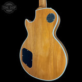 The back of an electric guitar with a natural wood finish, featuring a black pickguard and a circular black plate on the upper back, set against a solid black background.