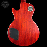 The back of a Gibson Les Paul Custom electric guitar with a glossy reddish-brown wood finish, featuring a black pickguard and a circular Gibson 'Deluxe Custom' badge on the upper right side, set against a solid black background.