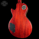 The back of a Gibson electric guitar with a glossy red cherry wood finish, featuring a circular silver 'Gibson Made in USA' badge and a black pickguard, shown against a solid black background.