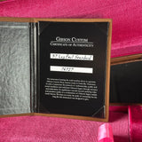 An open Gibson Custom Certificate of Authenticity booklet on a pink velvet surface, showing a black insert with white text and handwritten identification labels.