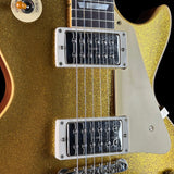 Close-up of a gold glitter electric guitar body with two black humbucker pickups, chrome hardware, and part of the neck visible against a dark background.