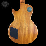 The back of a wooden electric guitar body, showing a warm honey-toned wood grain finish, a black pickguard, and a blue circular back plate badge, set against a solid black background.