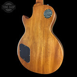 The back of a wooden electric guitar, showing the natural warm brown wood grain finish, a black pickguard, and a circular metal plate on a black background.