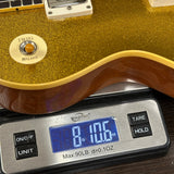 Close-up of a gold glitter electric guitar body resting on a digital scale displaying 8.105 oz, with a white volume/pitcher control and part of the brown wood neck visible.