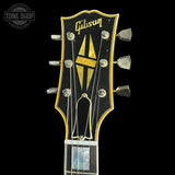 Close-up of a Gibson electric guitar headstock with a black face, gold outline, gold Gibson script logo, and a large gold and black geometric emblem. Six silver tuning pegs are visible against a black background.