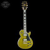 A Gibson Les Paul Custom electric guitar in a metallic gold finish, shown from the front against a solid black background. The guitar features a black pickguard, dual humbucker pickups, and a black-and-white block inlay fretboard.