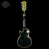 A vintage-style electric guitar shown from the back, featuring a black body with gold binding and a worn black finish, set against a solid black background.
