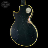 The back of a black Gibson electric guitar with a yellow binding edge, showing significant wear, scratches, and rust on the body, set against a solid black background.