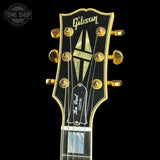 Close-up of a Gibson Les Paul Custom electric guitar headstock, featuring gold tuning pegs, a black headstock with gold trim, and a Gibson logo at the top, set against a solid black background.