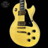 A gold-finish electric guitar with a black pickguard, black knobs, and gold hardware, shown against a solid black background with a Tone Shop watermark in the top left corner.