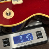 A close-up of a red electric guitar body resting on a digital scale displaying 9.45 oz, with the scale showing brand text 'WEIGHT Wx'.