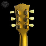 Close-up rear view of a guitar headstock in a metallic gold finish, showing six cream-colored tuning pegs and a black background. A Tone Shop watermark is visible in the upper left corner.