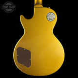 The back of a Gibson electric guitar in a metallic gold finish, shown against a solid black background. A Gibson logo badge is visible on the upper right, and a dark brown plate is mounted on the lower left.