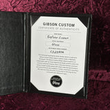 A Gibson Custom Certificate of Authenticity insert shown open on a dark textured surface, with a white label reading 'Gibson Custom Certificate of Authenticity' and instrument details including 'Explorer Custom' and 'Gloss'.