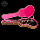 A black electric guitar with gold hardware resting inside an open hard case with a bright pink velvet interior and a brown wood-grain exterior, shown against a solid black background.