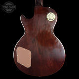 The back of a Gibson electric guitar with a glossy dark reddish-brown wood finish, featuring a circular gold-toned Gibson logo badge and a tan pickguard on a black background.