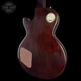 The back of a Gibson electric guitar with a glossy dark reddish-brown wood finish, featuring a circular gold Gibson logo badge and a gold pickguard surround, set against a solid black background.