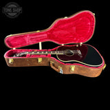 An acoustic guitar with a glossy black body and rosewood neck displayed inside an open vintage-style brown wooden hard case with bright red plush lining. The Tone Shop logo is visible in the top left corner.