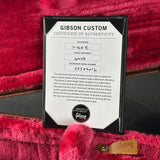 A Gibson Custom Certificate of Authenticity card displayed on a black frame, set against a bright pink plush velvet background with a small tan Gibson logo tag nearby.