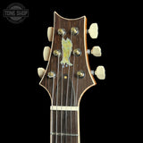 Close-up of the headstock of a six-string acoustic guitar against a solid black background. The headstock features a dark brown wood grain finish, cream-colored tuning pegs, and a central gold-toned logo badge.