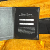 A black Gibson Custom Certificate of Authenticity booklet lies open on a yellow textured fabric background. The right page shows printed text and handwritten white label tapes over two lines, with the Gibson logo and 'Gibson Custom Certificate of Authenticity' visible.