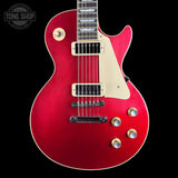 A red electric guitar with a cream pickguard and gold-toned hardware, shown against a solid black background. A Tone Shop logo is visible in the top-left corner.