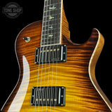 Close-up of a PRS electric guitar body showing a flamed maple top in a natural brown to golden yellow sunburst finish, with black humbucker pickups and gold hardware, set against a solid black background.