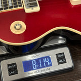 A close-up of a red electric guitar body resting on a digital scale, with the scale displaying 8.14 oz.