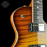 Close-up of a double-cutaway electric guitar body showing a natural wood flamed finish, control switch, pickups, and part of the fretboard on a black background.