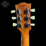 Close-up of the back of a guitar headstock made of warm brown wood, with six black tuning pegs and pale green/cream tuning keys, shown against a black background. A Tone Shop watermark is visible in the upper left.