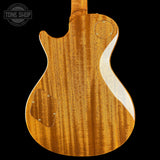 The back of an electric guitar with a rich, warm honey-amber wood finish and prominent grain patterns, shown against a solid black background. A circular Tone Shop watermark is visible in the top-left corner.