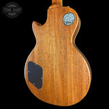 The back of a Gibson electric guitar with a natural brown wood finish, featuring a black pickguard and a circular Gibson logo badge on a black background.