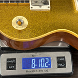 A close-up of a gold glitter electric guitar body resting on a digital scale displaying 8.12 oz.
